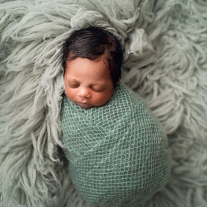 May include: A newborn baby is swaddled in a green knitted blanket and sleeping on a light green fluffy surface.