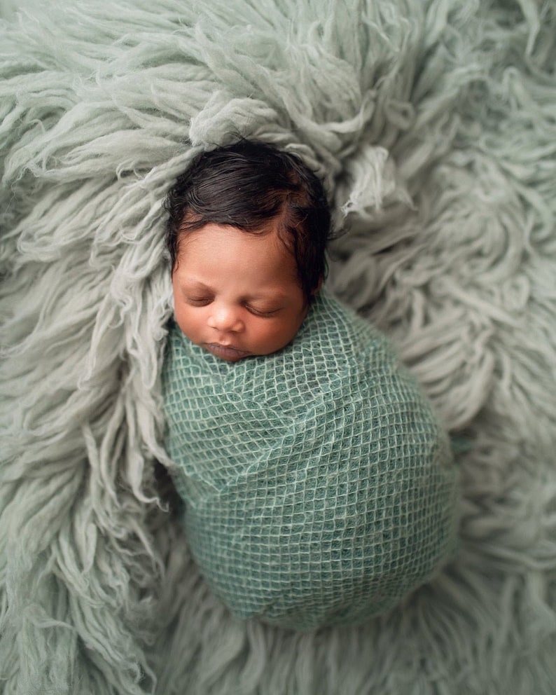 May include: A newborn baby is swaddled in a green knitted blanket and sleeping on a light green fluffy surface.