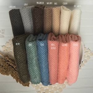 May include: A set of 14 knitted newborn photography props in various colors, including charcoal, gray, magnolia, walnut, oak, cream, off-white, olive, jade,  blue, rose blush, salmon, and blush. The props are arranged in a row and have a textured, honeycomb-like pattern.
