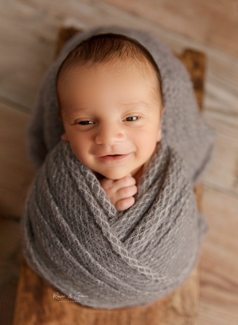 May include: A newborn baby wrapped in a gray knitted blanket, smiling up at the camera.
