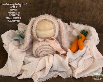 May include: A newborn photography prop set featuring a bunny bonnet, wrap, and two orange carrots. The bonnet and wrap are a soft, light pink color. The carrots have green tops. The set is designed for baby photoshoots.