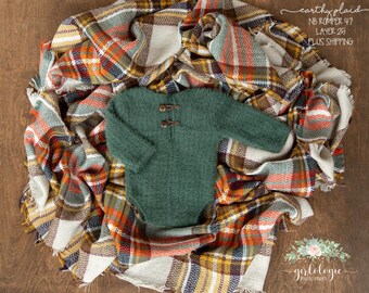 May include: A green newborn romper with wooden toggle closures is displayed on a plaid blanket with orange, brown, and cream colors. The blanket is draped on a wooden surface. The text "earthy plaid" and "NB Romper 47" is visible.