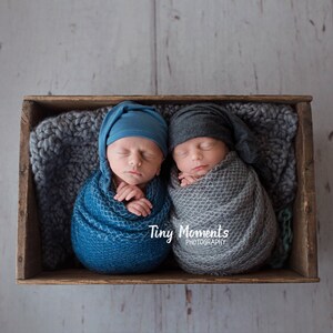 May include: Two newborn babies swaddled in blue and gray blankets, sleeping in a wooden crate with a gray knitted blanket. The babies are wearing blue and gray knit hats. The text "Tiny Moments Photography" is visible in the image.