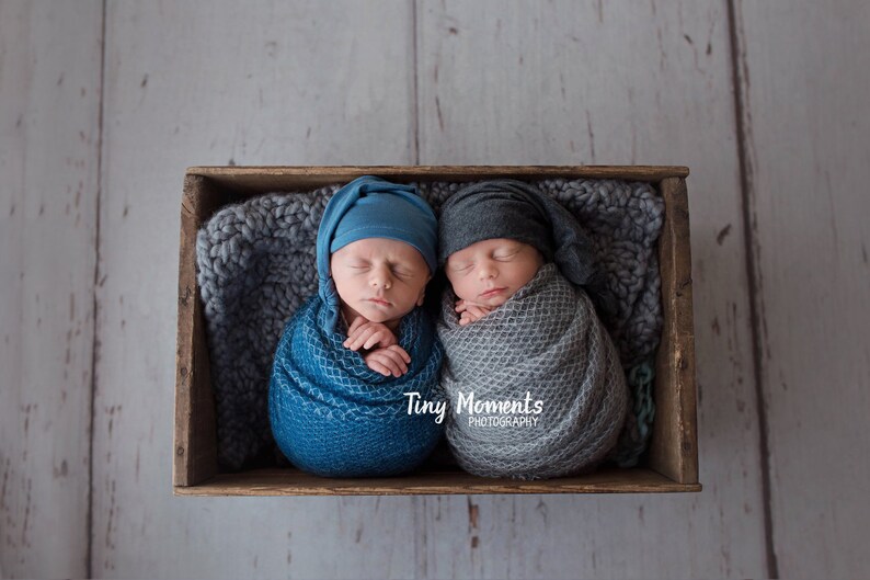 May include: Two newborn babies swaddled in blue and gray blankets, sleeping in a wooden crate with a gray knitted blanket. The babies are wearing blue and gray knit hats. The text "Tiny Moments Photography" is visible in the image.