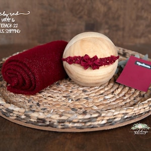 May include: A ruby red baby wrap and tieback set, with a wooden prop and a small card on a woven tray. The wrap is rolled up, and the tieback features a bow. The card is labeled "Madison".