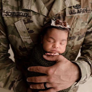 May include: A soldier in a U.S. Army uniform holds a sleeping baby wrapped in a green blanket. The soldier's name tag reads "Scarborough".