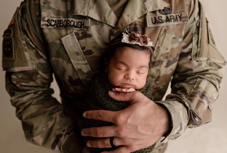 May include: A soldier in a U.S. Army uniform holds a sleeping baby wrapped in a green blanket. The soldier's name tag reads "Scarborough".