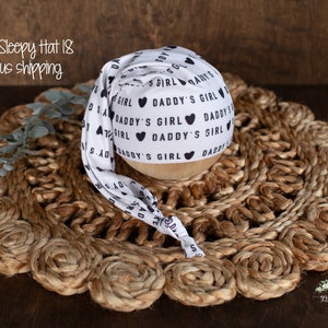 Puede incluir: Sombrero de dormir blanco para recién nacido con texto negro que dice "Daddy's Girl" y corazones negros. El sombrero está sobre una estera de paja tejida.