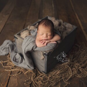 May include: A newborn baby is sleeping peacefully in a wooden crate lined with a gray blanket and beige fabric. The crate is sitting on a wooden floor.
