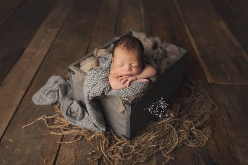 May include: A newborn baby is sleeping peacefully in a wooden crate lined with a gray blanket and beige fabric. The crate is sitting on a wooden floor.