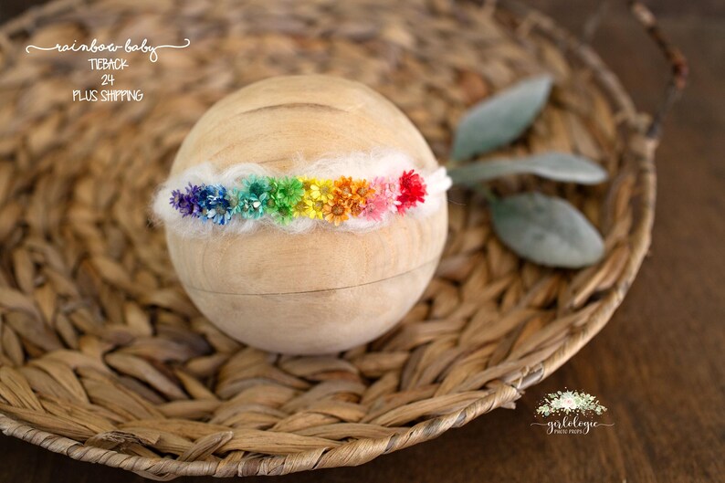 May include: A rainbow flower headband with white felt and colourful flowers. The headband is on a woven basket.