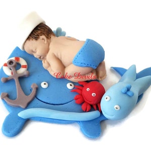 May include: A blue whale cake topper with a sleeping baby wearing a sailor hat and blue shorts. The whale has a red crab and an anchor on it.