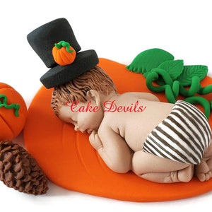 May include: A fondant baby figurine wearing a black top hat with a small orange pumpkin on top, lying on a large orange pumpkin with green leaves and a vine. The baby is wearing a black and white striped diaper. There is a small orange pumpkin and a brown pine cone on the side.