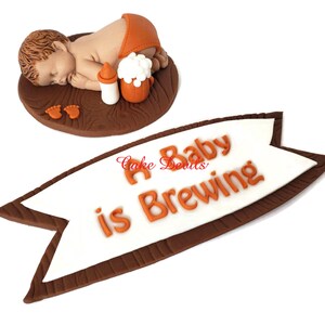 May include: A brown and white cake topper with a baby sleeping on a wooden board. The baby is holding a bottle and a beer mug. A banner reads "A Baby is Brewing".