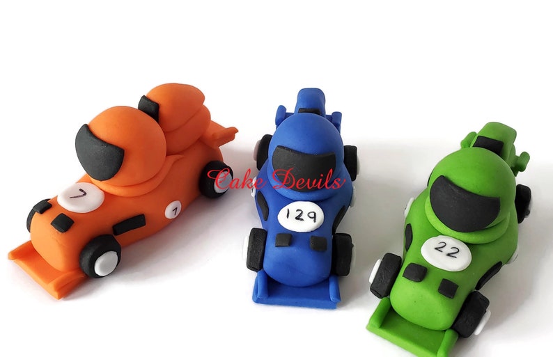 May include: Three fondant race car toppers, one orange, one blue, and one green, with black racing helmets and white numbers on the sides. The orange car has the number 7, the blue car has the number 129, and the green car has the number 22.