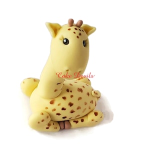Puede incluir: Un adorno para pastel de jirafa de fondant amarillo y marr&oacute;n con manchas marrones. La jirafa tiene un cuello largo y una cabeza peque&ntilde;a con ojos marrones. La jirafa est&aacute; sentada sobre sus patas traseras con sus patas delanteras metidas.