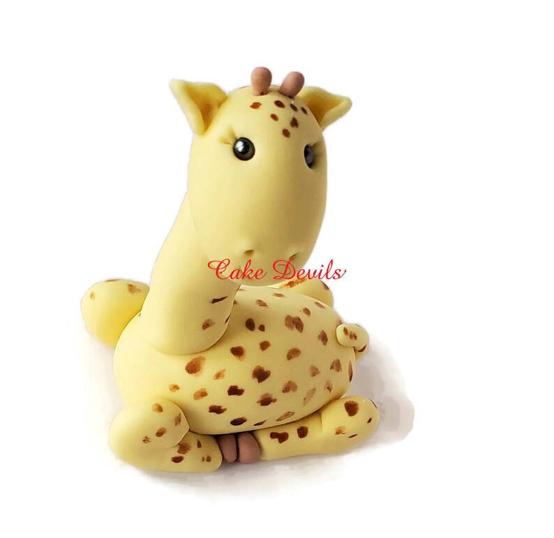 Puede incluir: Un adorno para pastel de jirafa de fondant amarillo y marr&oacute;n con manchas marrones. La jirafa tiene un cuello largo y una cabeza peque&ntilde;a con ojos marrones. La jirafa est&aacute; sentada sobre sus patas traseras con sus patas delanteras metidas.