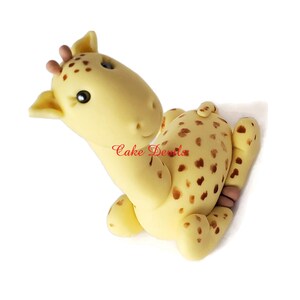 Puede incluir: Un adorno para pastel de fondant de jirafa amarillo y marr&oacute;n con manchas marrones. La jirafa est&aacute; sentada con la cabeza girada hacia un lado y los ojos cerrados.