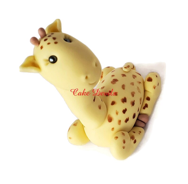 Puede incluir: Un adorno para pastel de fondant de jirafa amarillo y marr&oacute;n con manchas marrones. La jirafa est&aacute; sentada con la cabeza girada hacia un lado y los ojos cerrados.