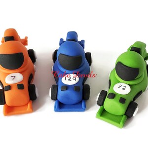 May include: Three fondant race car toppers, one orange, one blue, and one green. Each car has a black visor, a white number, and black wheels.