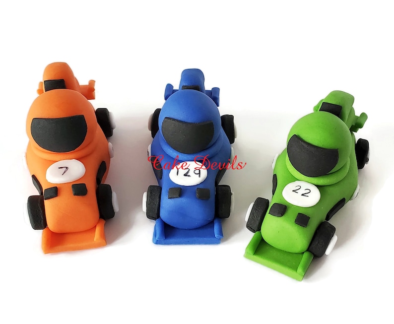 May include: Three fondant race car toppers, one orange, one blue, and one green. Each car has a black visor, a white number, and black wheels.