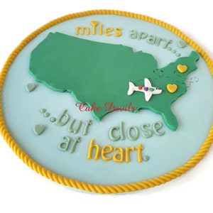 May include: A round, light blue cake decorated with a green map of the United States, a white airplane, and the text "miles apart...but close at heart." The border is yellow rope-like icing. Small heart shapes are scattered around the map.