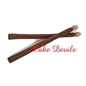 May include: Two brown wooden drumsticks with beige tips. The drumsticks are crossed over each other. The text "Cake Devils" is written in red cursive below the drumsticks.