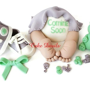 May include: A baby shower cake topper featuring a baby's bottom with a gray blanket and the words "Coming Soon" in green. The topper also includes a pair of gray and white baby shoes, a green bow, a green teddy bear, and other baby-themed decorations.