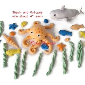 May include: A set of 15 fondant cake decorations featuring an orange octopus, a gray shark, and various sea life elements, including yellow fish, seashells, and starfish. The shark and octopus are approximately 4 inches tall.