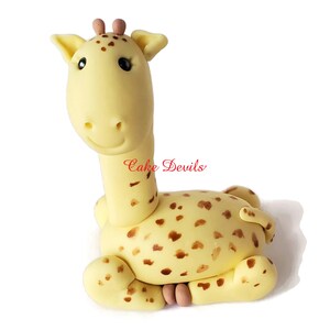 Puede incluir: Un adorno para pastel de jirafa de fondant amarillo con manchas marrones. La jirafa est&aacute; sentada con las piernas metidas debajo del cuerpo. La jirafa tiene la nariz y las orejas rosas.