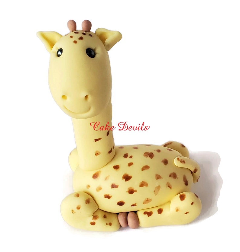 Puede incluir: Un adorno para pastel de jirafa de fondant amarillo con manchas marrones. La jirafa est&aacute; sentada con las piernas metidas debajo del cuerpo. La jirafa tiene la nariz y las orejas rosas.
