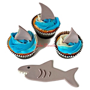 May include: Three blue and white frosted cupcakes with gray fondant shark fin toppers. A separate gray fondant shark cutout is also included.