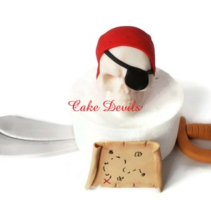 May include: A white cake with a red and white pirate skull topper, a brown sword, and a brown treasure map with an X marking the spot.