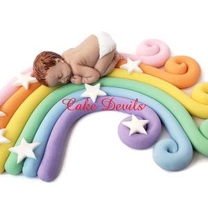 May include: A sleeping baby figurine on a rainbow cake topper with white stars. The rainbow is made of fondant and has a light pink, yellow, green, light blue, and purple color scheme. The cake topper is decorated with the text "Cake Devils".