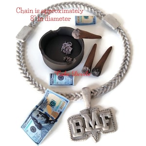 May include: A silver chain necklace with a pendant that says "BMF". The chain is 8 inches in diameter and is decorated with rolled-up dollar bills, cigars, and an ashtray.