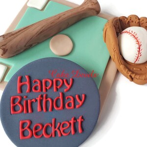 May include: A baseball themed cake topper with a brown bat, a baseball glove with a white baseball, and a blue circle with the text "Happy Birthday Beckett" in red.