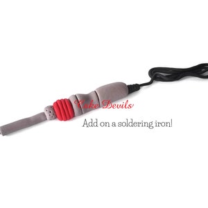 May include: A gray and red fondant soldering iron with a black cord. The text "Cake Devils" and "Add on a soldering iron!" is written below the iron.