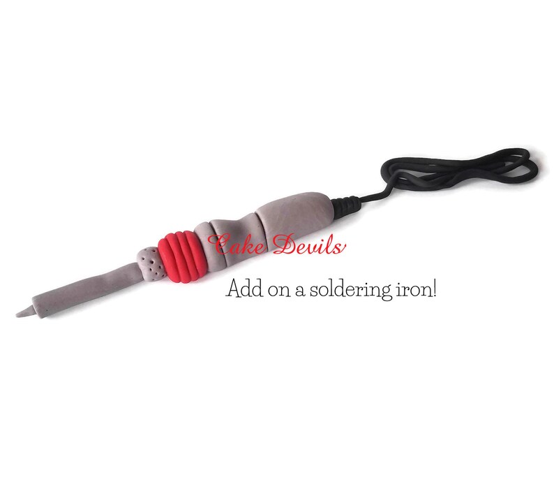 May include: A gray and red fondant soldering iron with a black cord. The text "Cake Devils" and "Add on a soldering iron!" is written below the iron.