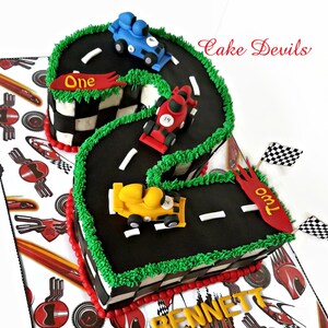 May include: A black and green race track shaped like the number two with three toy race cars on it. The cars are red, yellow, and blue. The cake is decorated with red and white checkered flags and the words "One" and "Two" on the sides. The cake is on a white background with a red and white checkered pattern.