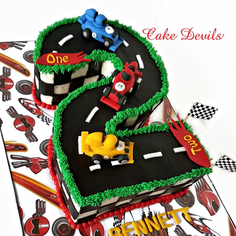 May include: A black and green race track shaped like the number two with three toy race cars on it. The cars are red, yellow, and blue. The cake is decorated with red and white checkered flags and the words "One" and "Two" on the sides. The cake is on a white background with a red and white checkered pattern.