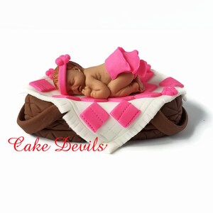 May include: A pink and white fondant baby girl cake topper. The baby is sleeping on a pink and white blanket in a brown basket. The cake topper is decorated with pink flowers.