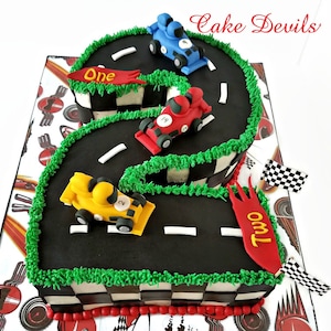 May include: A black and green race track shaped like the number two with three toy race cars on it. The cars are red, blue, and yellow. The cake is decorated with a checkered flag pattern and the words "One" and "Two" on the track.