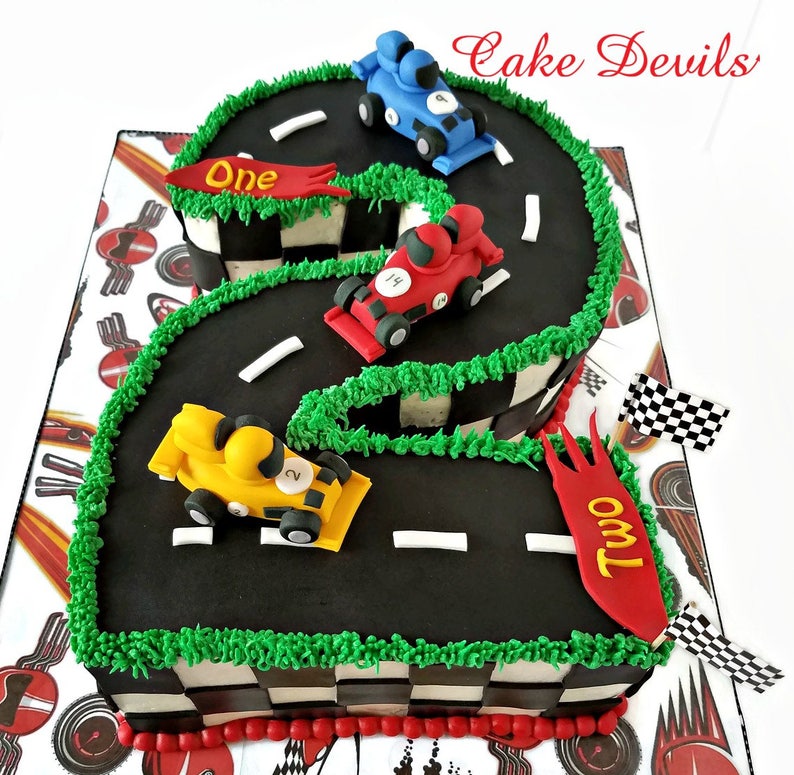 May include: A black and green race track shaped like the number two with three toy race cars on it. The cars are red, blue, and yellow. The cake is decorated with a checkered flag pattern and the words "One" and "Two" on the track.