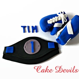 May include: A blue and white boxing glove cake topper with a black and blue championship belt with the number "1" on it. The name "TIM" is spelled out in blue icing. The text "Cake Devils" is written in red cursive.