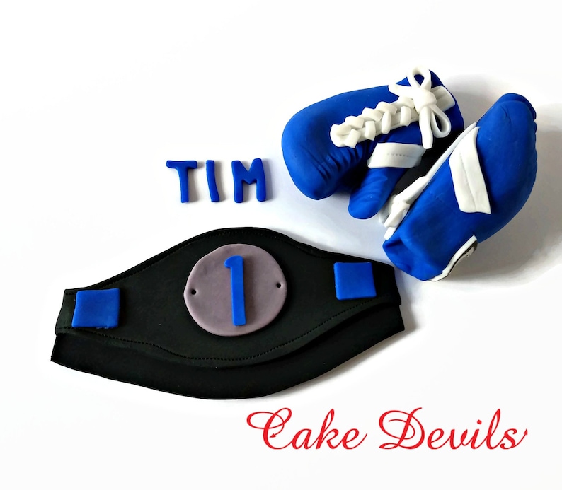 May include: A blue and white boxing glove cake topper with a black and blue championship belt with the number "1" on it. The name "TIM" is spelled out in blue icing. The text "Cake Devils" is written in red cursive.