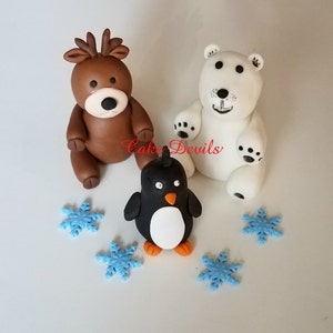 May include: A set of three fondant cake toppers featuring a brown reindeer, a white polar bear, and a black and white penguin. The penguin is standing on a white surface with blue snowflake sprinkles.