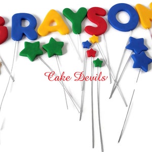 May include: A cake topper set featuring the name "GRAYSON" in colorful letters and stars. The letters are red, blue, yellow, and green. The stars are yellow, green, red, and blue. The letters and stars are attached to silver wires.