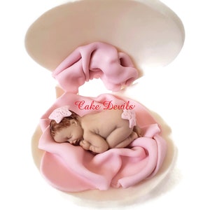 May include: A pink fondant baby doll sleeping in a pink blanket inside a white fondant seashell. The doll has a pink bow on its head. The text "Cake Devils" is visible on the seashell.