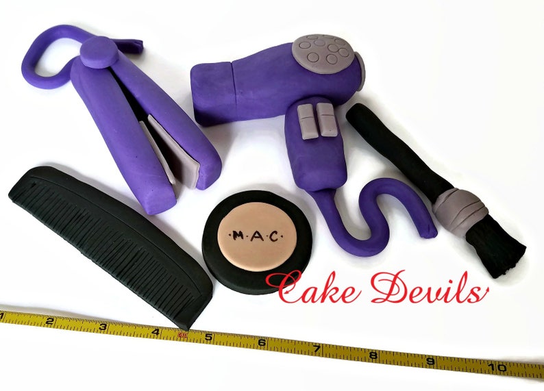 May include: Purple hair styling tools made of fondant, including a hairdryer, curling iron, comb, and a makeup compact with the text "M.A.C."