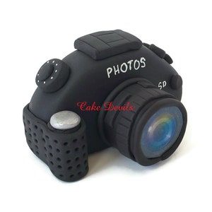 May include: Black camera cake topper with the word "PHOTOS" written on the top. The camera has a lens with a rainbow effect and a silver button on the side.